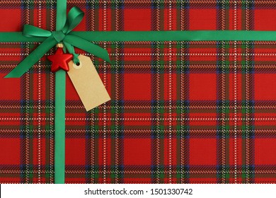 Christmas Present, Gift Close-up. Green Satin Ribbon, Bow With Red Ornament And Empty Tag On Scottish Tartan Plaid Paper.  