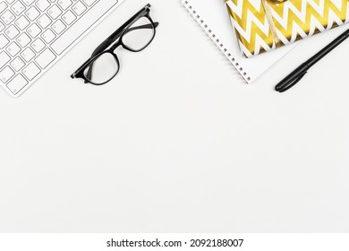 Christmas Present Wrapped In Golden Paper, Keyboard, Black Eyeglasses, Pen, Notepad On White Background. Office Work Desk. Business Celebration Layout. Secret Santa Concept. Copy Space, Flat Lay.