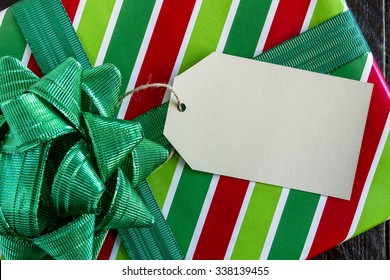 Christmas Present Wrapped In Red And Green Striped Wrapping Paper With Shiny Green Bow And Blank Tag Sitting On Wooden Table