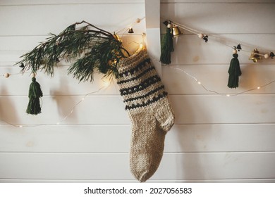 Christmas Rustic Stocking With Fir Branch And Stylish Garland With Bells And Trees On White Wooden Wall With Christmas Lights. Festive Decorated Atmospheric Scandinavian Room. Modern Handmade Decor