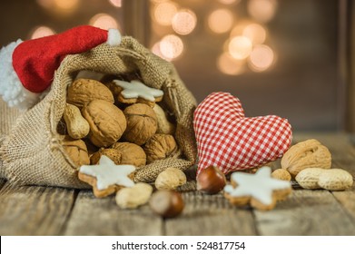 Christmas sack, advent season, decoration with nuts, cookies, red heart and Santa hat on wooden table with twinkling lights background.
