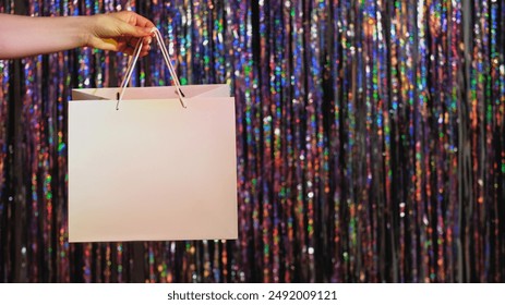 Christmas sale. Holiday shopping. Black Friday offer. Woman hand holding brand mockup paper bag with empty space on defocused glitter iridescent tinsel curtain festive background. - Powered by Shutterstock