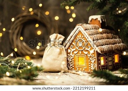 Christmas Santa Bag with Gifts next to Gingerbread House. White Burlap Sack with Xmas Presents over shining Lights Background. Ginger Cake with Icing. Winter Holiday Home Ornament