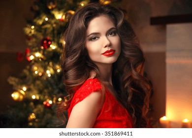 Christmas Santa. Beautiful Smiling Woman Model. Makeup. Healthy Long Hair Style. Elegant Lady In Red Dress Over Christmas Tree Lights Background. Happy New Year.