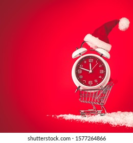 Christmas Santa Hat On Red Clock, Balanced In Shopping Cart. Last Minute Christmas Shopping Concept With Blank Empty Space For Text.