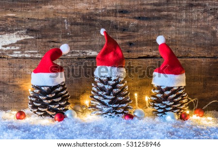 Christmas season, rustic decoration, Santa hats on pine cones with lights at snow and brown wooden background.