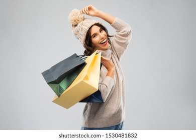 Christmas, Seasonal Sale And Consumerism Concept - Happy Smiling Young Woman In Knitted Winter Hat And Sweater With Shopping Bags Over Grey Background