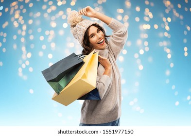 Christmas, Seasonal Sale And Consumerism Concept - Happy Smiling Young Woman In Knitted Winter Hat And Sweater With Shopping Bags Over Festive Lights On Blue Background