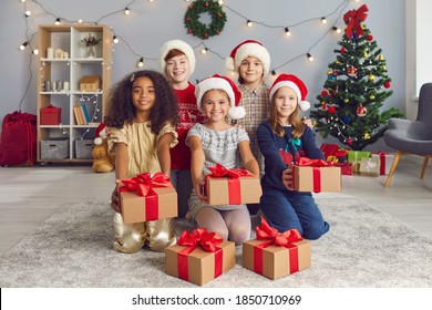 Christmas Is For Sharing. Group Of Happy Smiling Diverse Little Children In Santa Caps Looking At Camera And Holding Out Craft Cardboard Gift Boxes Tied With Red Bows, As If Giving Presents To You