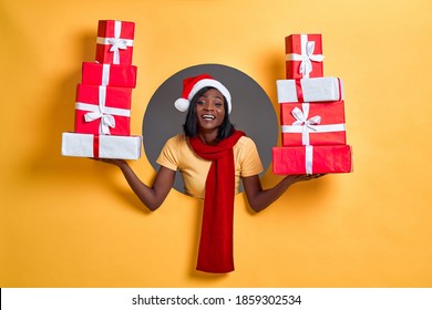 Christmas shopping African American woman holding many gifts in her arms wearing santa hat and red scarf in a round circle hole in orange background.