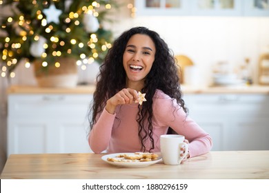 Christmas Snack. Joyful brunette woman enjoying holiday cookies and hot chocolate in kitchen, eating tasty xmas decorated biscuits and drinking coffee, celebrating winter holidays at home, copy space