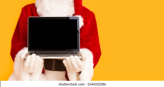 Christmas Snow Flowing Of Gadgets. Santa Claus Showing Modern Laptop With Black Blank Screen On Orange Background, Panorama, Copy Space