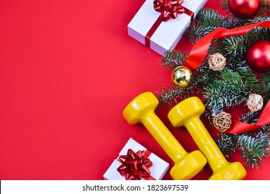 Christmas Sports Equipment. Dumbbells, Red Ribbon, Fir Branch And White Gift Box With A Red Bow On A Red Background. Healthy Lifestyle And Sport Concept. View From Above. Copy Space.