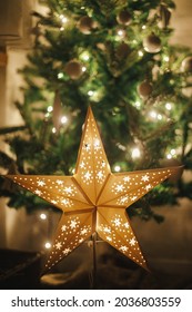 Christmas Star On Background Of Christmas Tree In Lights In Evening Room. Big Paper Star Garland Glowing In Festive Decorated Scandinavian Room. Atmospheric Christmas Eve. Happy Holidays