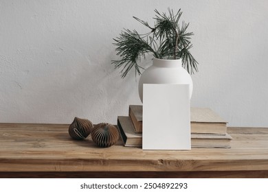 Christmas stationery. Blank greeting card, invitation mockup. Vintage wooden bench, table. Modern vase with pine tree branches. Paper christmas balls, white wall background. Scandinavian interior.