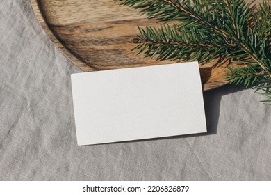 Christmas stationery. Closeup of blank business card, invitation mockup. Green spruce tree branch on wooden tray in sunlight. Grey linen tablecloth. Festive winter still life template. Flat lay, top.