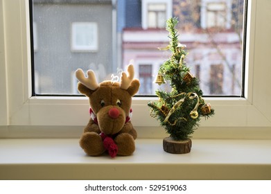 Christmas Still Life With Small Christmas Tree And Funny Puppet Toy Reindeer Rudolf With Red Nose On Windowsill