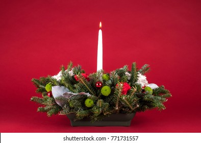 Christmas Table Arrangement Or Centerpiece With Flowers, Evergreen Branches And White Lit Candle On Red