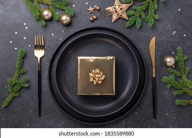 Christmas table setting. Black ceramic plate with golden gift box, fir tree branch and accessories on stone background. Gold decoration - Image - Powered by Shutterstock