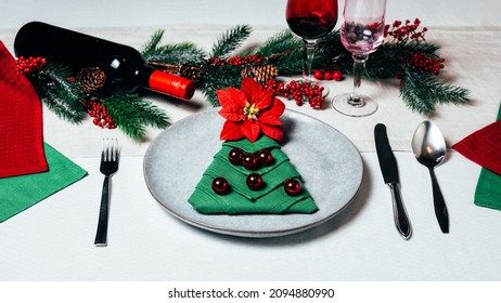Christmas table setting with ceramic dishware, silver tableware and green festive tree napkin folds and poinsettia on white cloth. Top view.