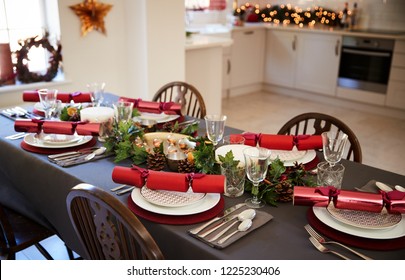 Christmas Table Setting With Christmas Crackers Arranged On Plates In A Dining Room, With Kitchen In The Background