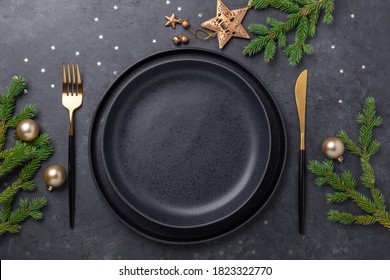 Christmas Table Setting With Empty Black Ceramic Plate, Fir Tree Branch And Gold Accessories On Black Stone Background. Top View. Copy Space - Image