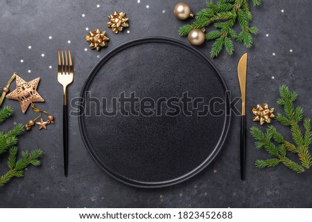 Christmas table setting with empty black ceramic plate, fir tree branch and gold accessories on black stone background. Top view. Copy space - Image