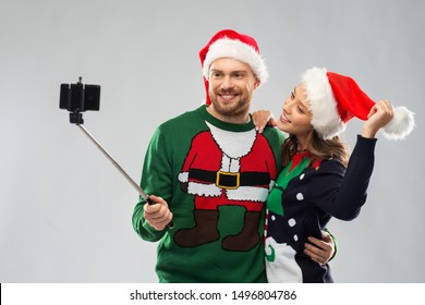 Christmas, Technology And Holidays Concept - Happy Couple In Santa Hats Taking Picture By Smartphone On Selfie Stick At Ugly Sweater Party