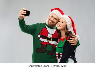 Christmas, Technology And Holidays Concept - Happy Couple In Santa Hats Taking Selfie By Smartphone At Ugly Sweater Party