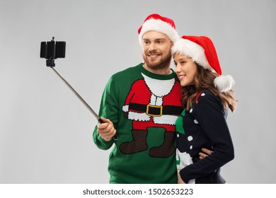 Christmas, Technology And Holidays Concept - Happy Couple In Santa Hats Taking Picture By Smartphone On Selfie Stick At Ugly Sweater Party