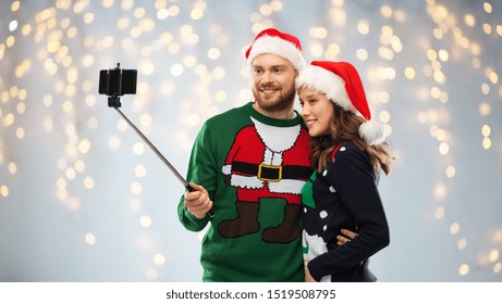 Christmas, Technology And Holidays Concept - Happy Couple In Santa Hats Taking Picture By Smartphone On Selfie Stick At Ugly Sweater Party Over Festive Lights Background