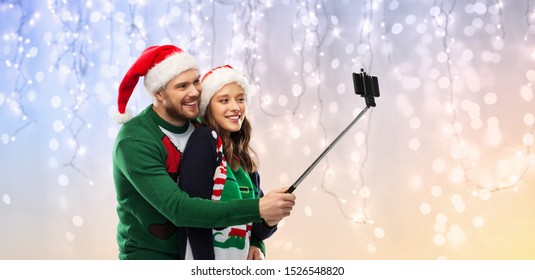 Christmas, Technology And Holidays Concept - Happy Couple In Santa Hats Taking Picture By Smartphone On Selfie Stick At Ugly Sweater Party Over Festive Lights Background