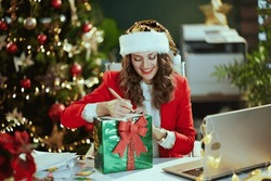 Christmas Time. Happy Elegant Middle Aged Business Woman In Santa Hat And Red Jacket With Laptop And Present Box Singing Postcard In Modern Green Office With Christmas Tree.