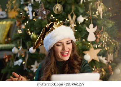 Christmas Time. Happy Trendy Housewife With Santa Hat In Green Dress Having Web Cam Meeting On A Tablet PC Near Christmas Tree In The Modern House.