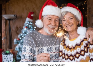 Christmas time. Portrait of blurred smiling senior couple in Santa hat holding sparkler lights at home. Celebration Christmas and new year concept - Powered by Shutterstock