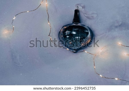 Christmas tree dark blue toy lies on white shiny snow surrounded by a garland with warm light of lanterns in evening light, view from above. Winter background with snow with bright festive decor.