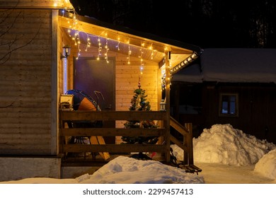 Christmas tree stands by entrance of wooden residential house decorated with Christmas lights garlands at night. Soft focus.  Winter holidays building decoration theme. - Powered by Shutterstock