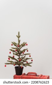 The Christmas Tree Stands On A Red Suitcase. New Year Decorations On A White Wall. Green Spruce With Red Toys Next To A Colored Case