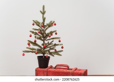 The Christmas Tree Stands On A Red Suitcase. New Year Decorations On A White Wall. Green Spruce With Red Toys Next To A Colored Case