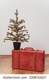 The Christmas Tree Stands On A Red Suitcase. New Year Decorations On A White Wall. Green Spruce With Red Toys Next To A Colored Case