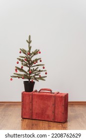 The Christmas Tree Stands On A Red Suitcase. New Year Decorations On A White Wall. Green Spruce With Red Toys Next To A Colored Case