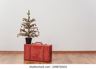 The Christmas Tree Stands On A Red Suitcase. New Year Decorations On A White Wall. Green Spruce With Red Toys Next To A Colored Case