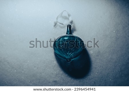 A Christmas tree toy in the form of a glass drop of dark blue color lies on white shiny snow in the evening light, top view. A symbol of winter holidays, joy and Christmas magic.