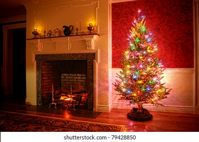 Christmas Tree With Vintage Multicolor Lights In An Old Fashioned Traditional Interior With Brick Fireplace And Warm Burning Fire Log In Hearth