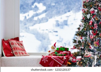 Christmas Tree At Window With View To Snow And Swiss Alps Mountains In Winter. Decorated Living Room With Xmas Gifts And Presents For Kids, Pillows And Toys. Family Home Seasonal Interior Decoration.