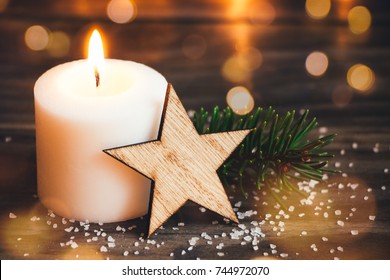 Christmas White Candle With A Wooden Star, Fir Branch And Lights Bokeh At The Background