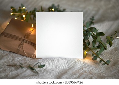 Christmas Winter Empty 5x5 Square Card Mockup With Gift, Eucaliptus Leaves And Light Garland On A Knitted Beige Background, Element For Invitation, Thank You O Greeting Card. Christmas Cozy Background