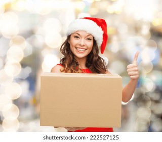 Christmas, Winter Holidays, Delivery, Gesture And People Concept - Smiling Woman In Santa Helper Hat With Parcel Box Showing Thumbs Up Over Lights Background