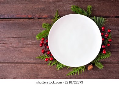 Christmas or winter table setting mock up. Blank white plate with frame of evergreen branches and red berries. Top view on a dark wood background. Copy space. - Powered by Shutterstock