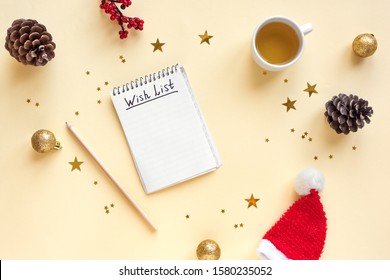 Christmas Wish List written on paper with cup of tea, pencil and Christmas festive decor, flat lay on yellow pastel background. New Year's wishing and planning concept, top view.